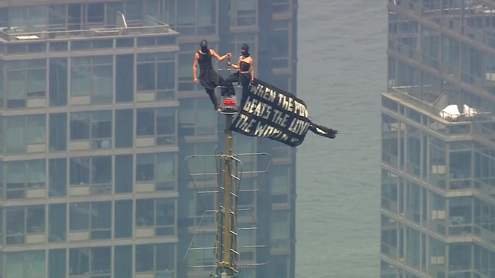 Angela Nikolau and Ivan Kuznetsov standing on the Empire State Building antenna in New York City on July 1, 2026, displaying a black peace banner