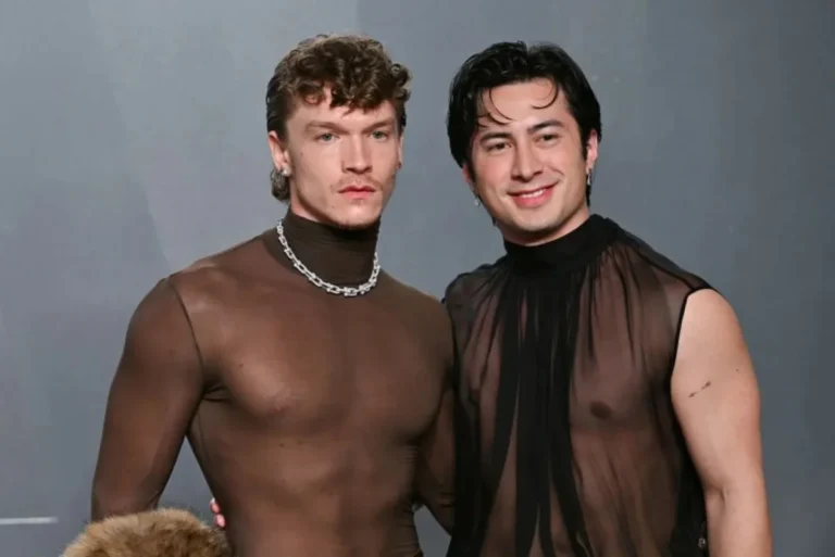 Two male leads in dark tuxedos standing face-to-face against a warm-toned blurred city background.