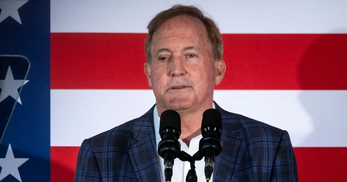 Image: Texas Attorney General Ken Paxton stands in front of a mic and a U.S. flag backdrop.