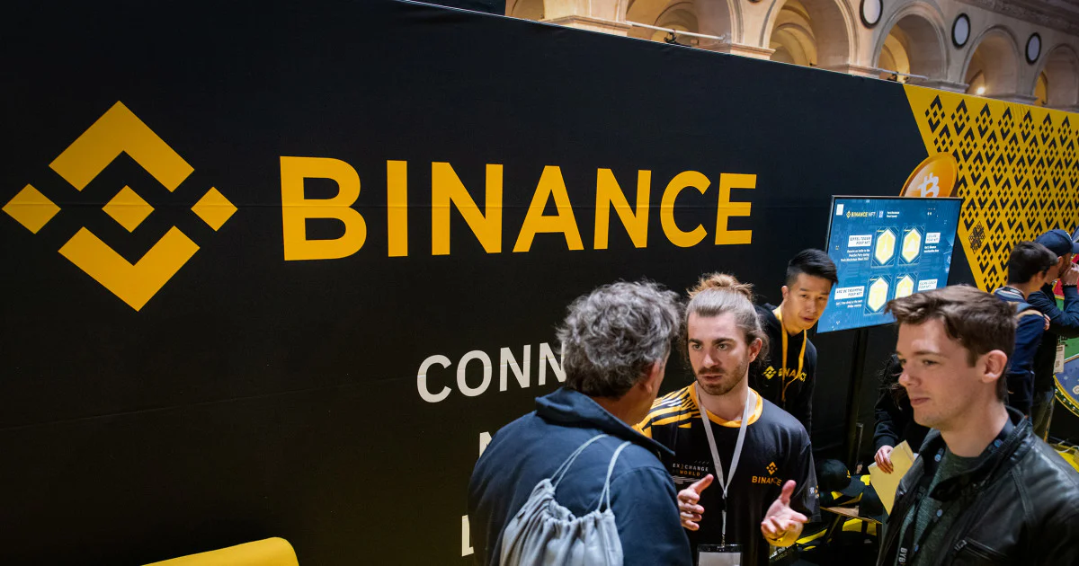 Binance cryptocurrency exchange logo displayed on a smartphone beside digital asset trading monitors during coverage of the October 2026 security breach